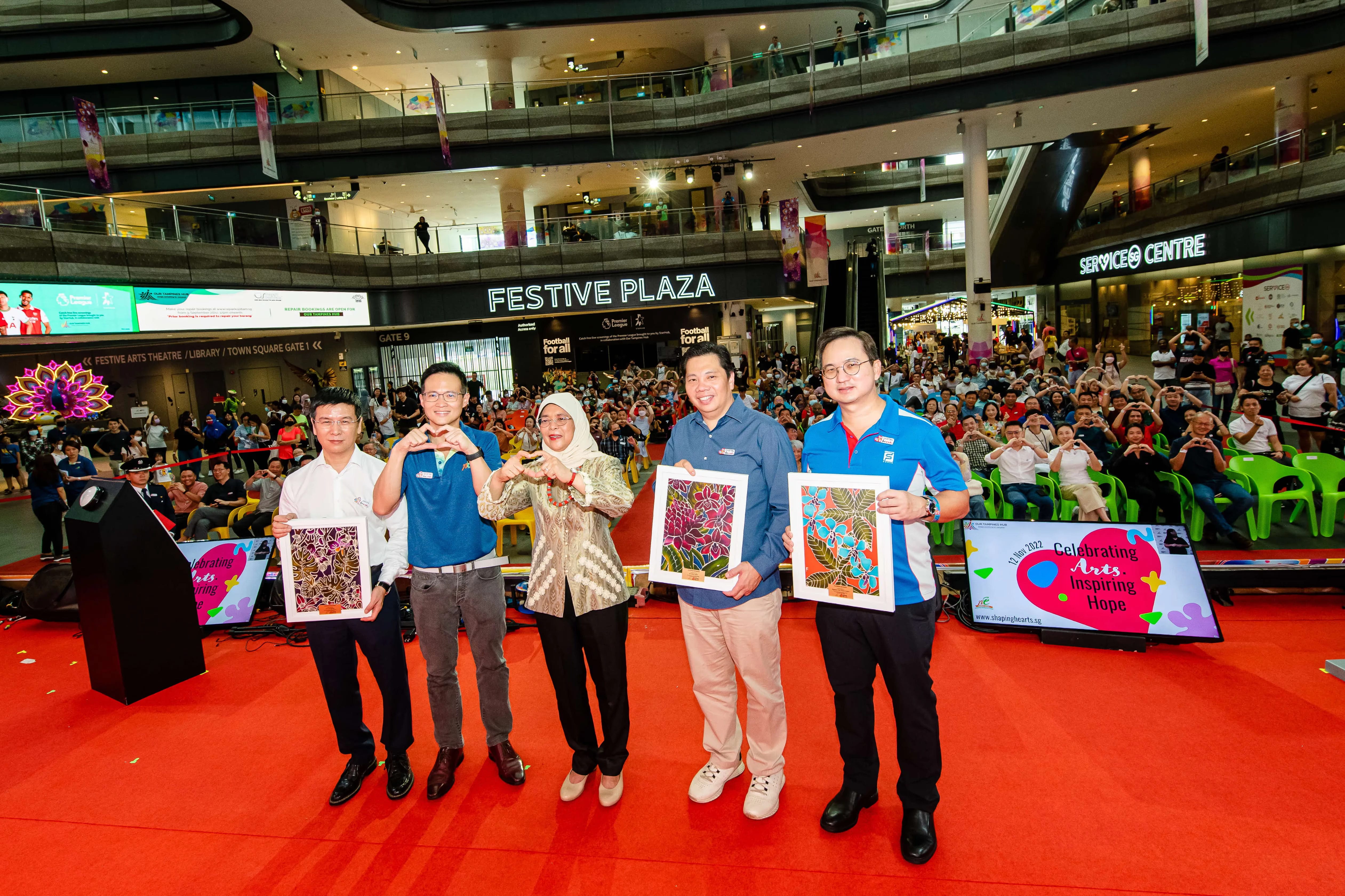 Five people stand on a red stage holding artwork, with a large crowd seated behind them in Festive Plaza.